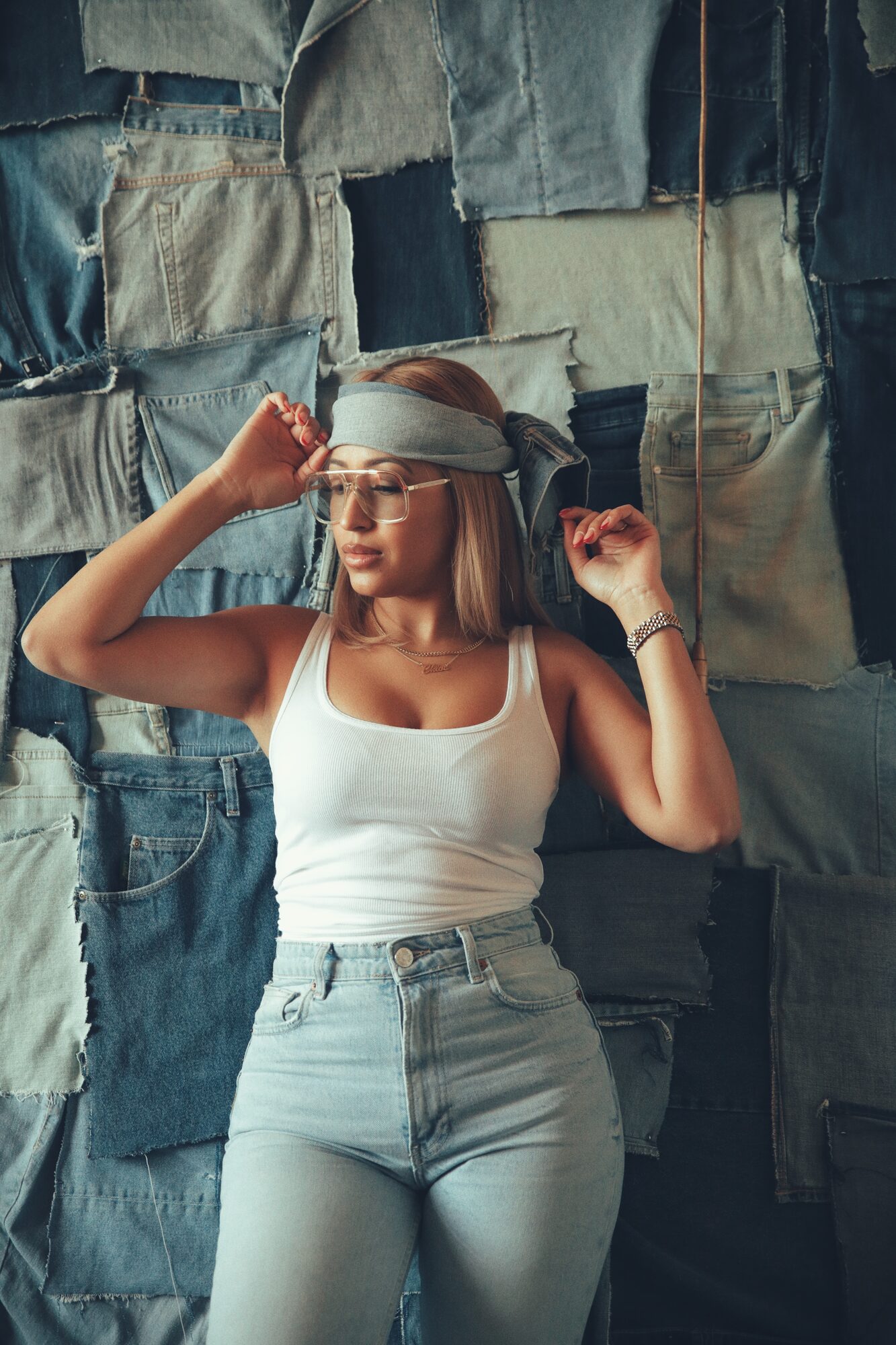 Woman in a white tank top and glasses adjusting a denim headband against a patchwork denim wall backdrop.