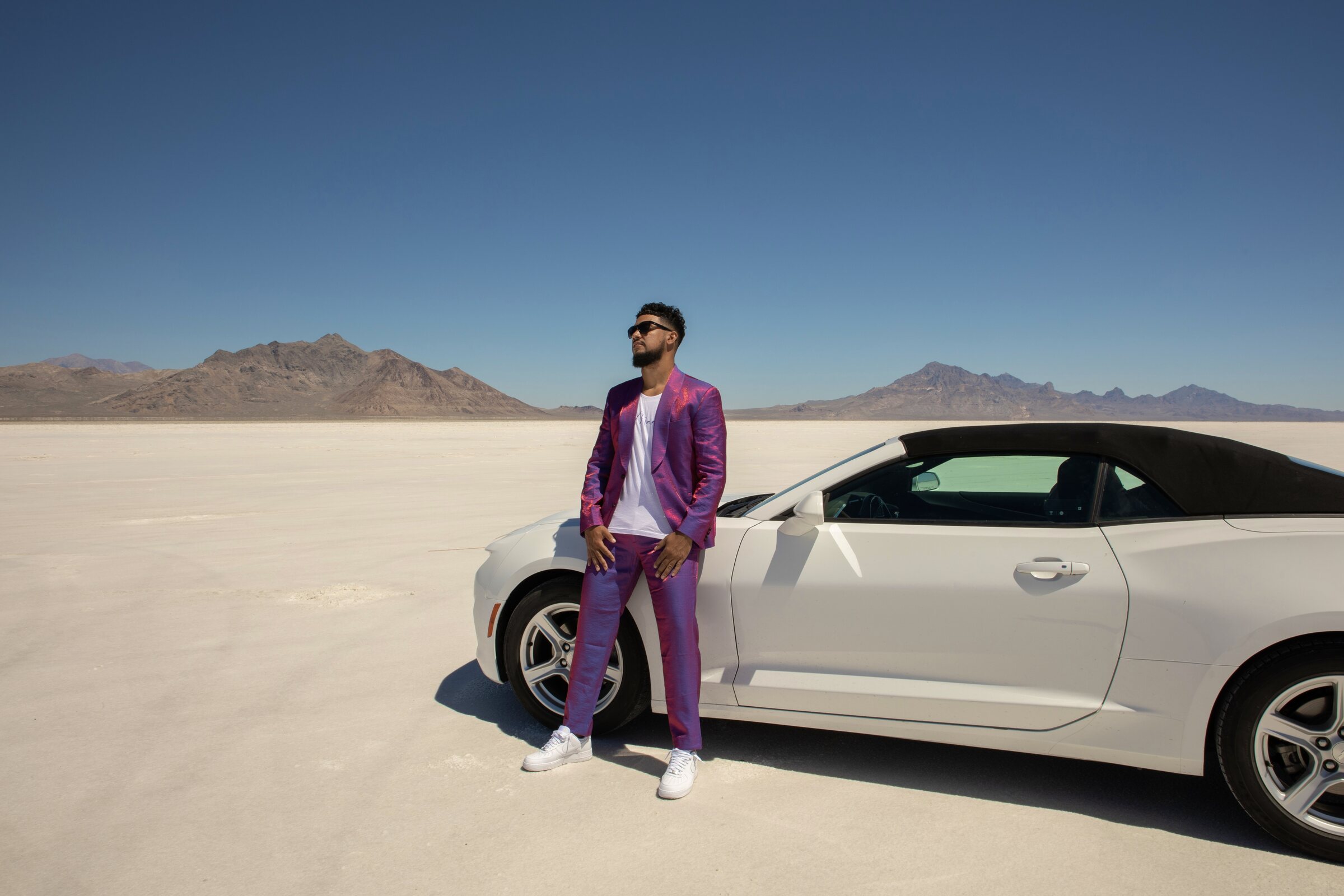 Model in an iridescent magenta suit leaning against a white convertible on the Bonneville Salt Flats, mountains on the horizon.