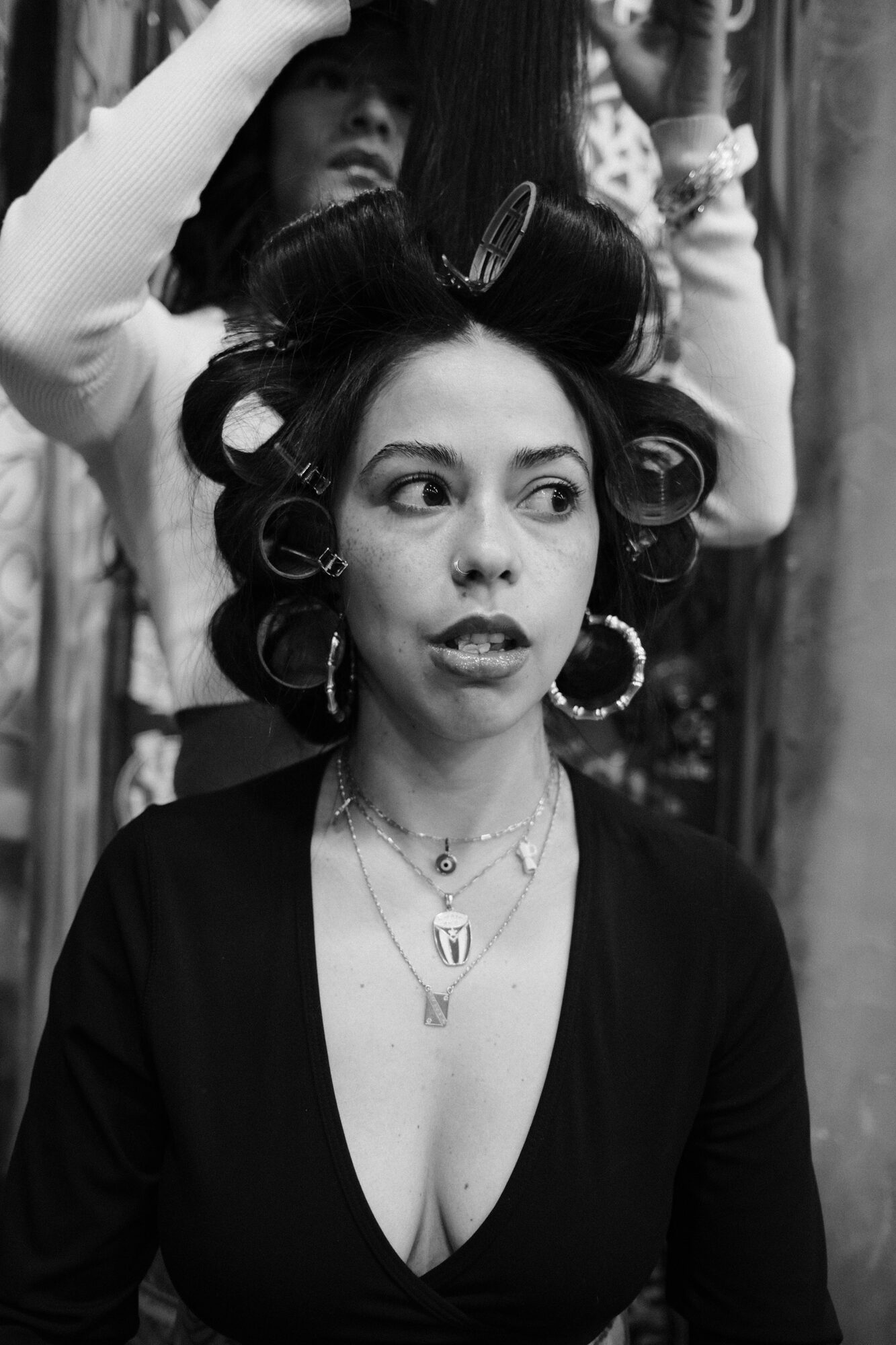 Black and white backstage candid of a woman with hair in curlers and hoop earrings, mid-conversation.