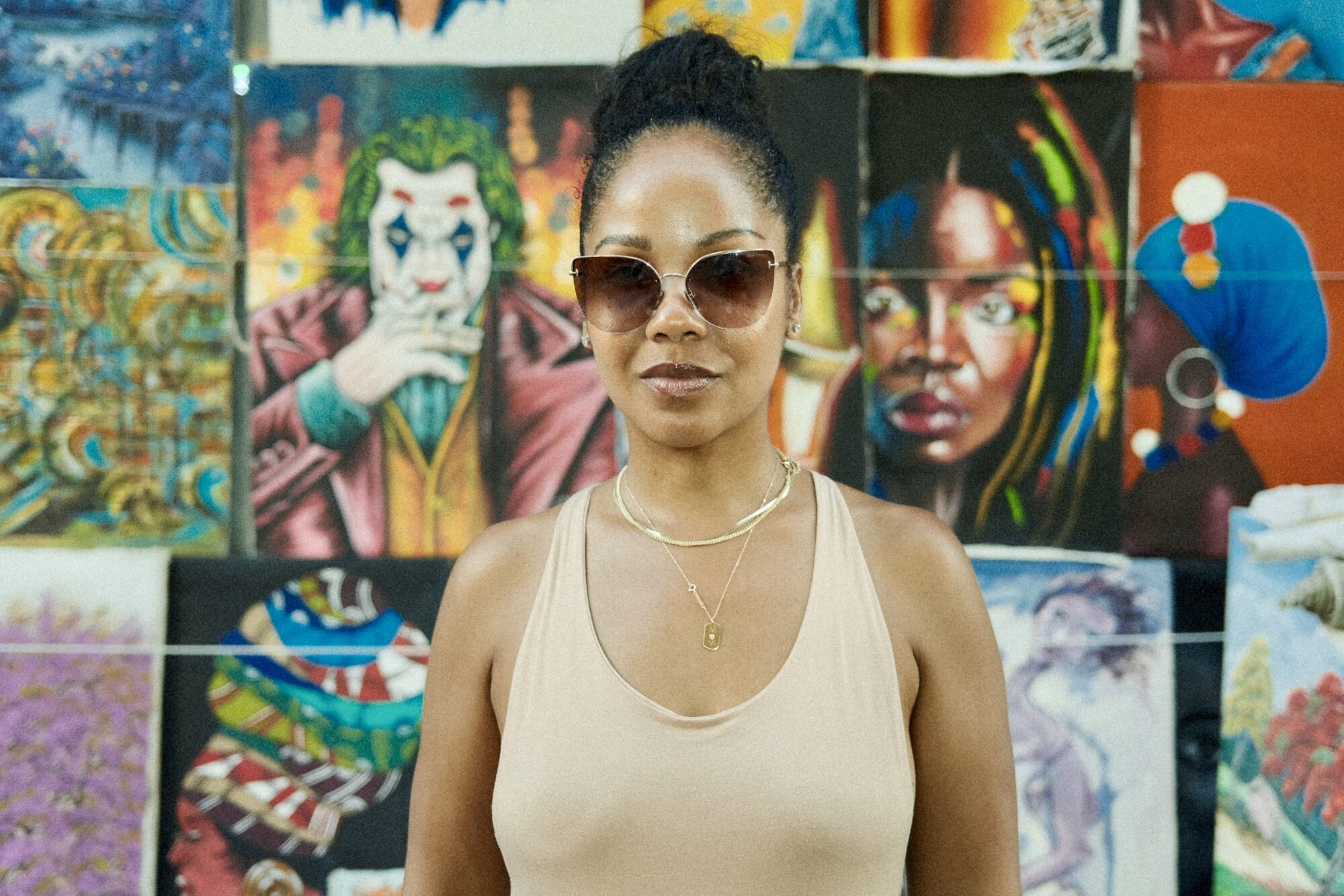 Portrait of a woman in oversized sunglasses and a tan tank top standing in front of a colorful street art mural wall.