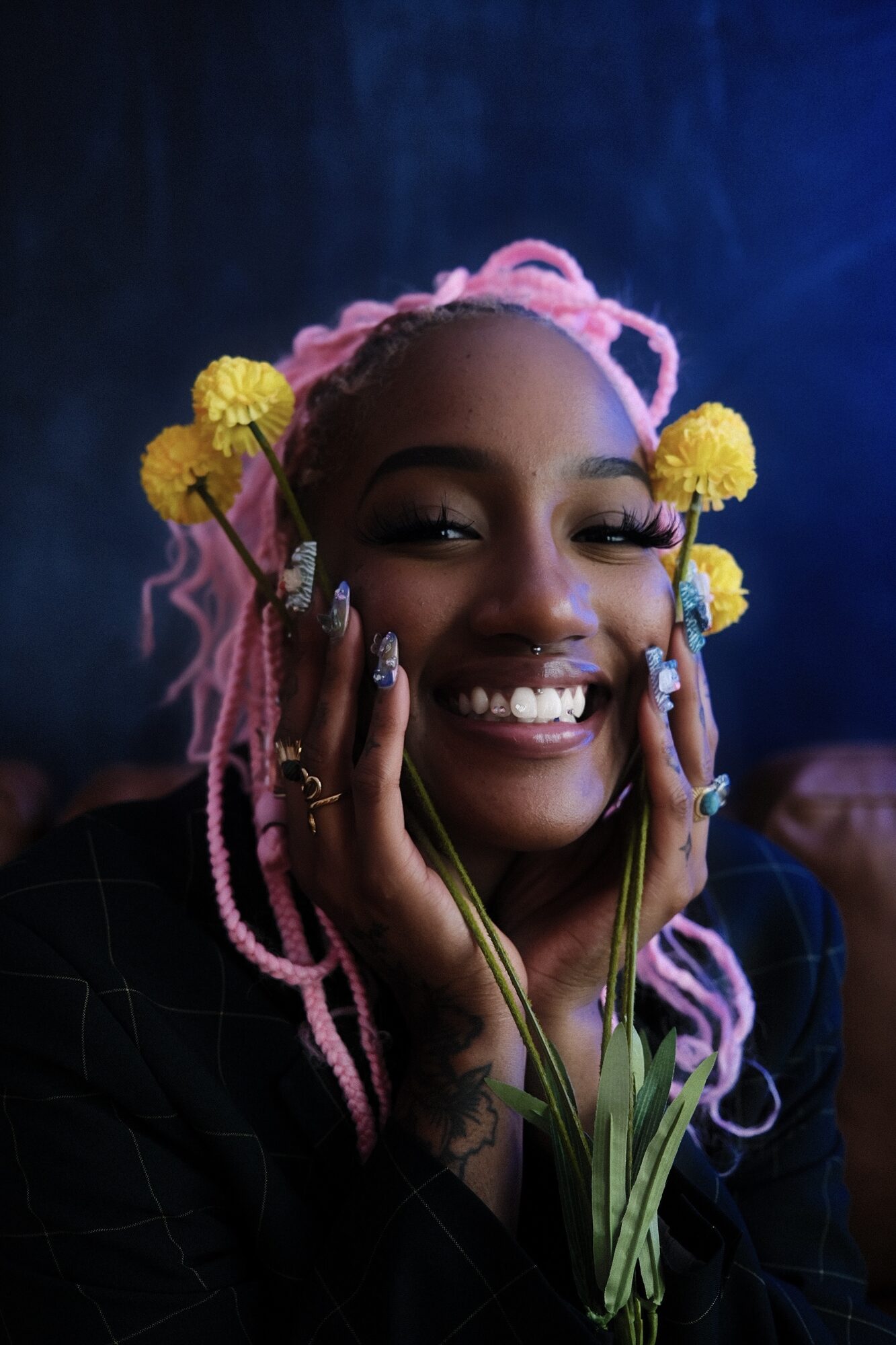 Smiling portrait of a woman with pink braided hair and yellow flowers framing her face, deep blue background.