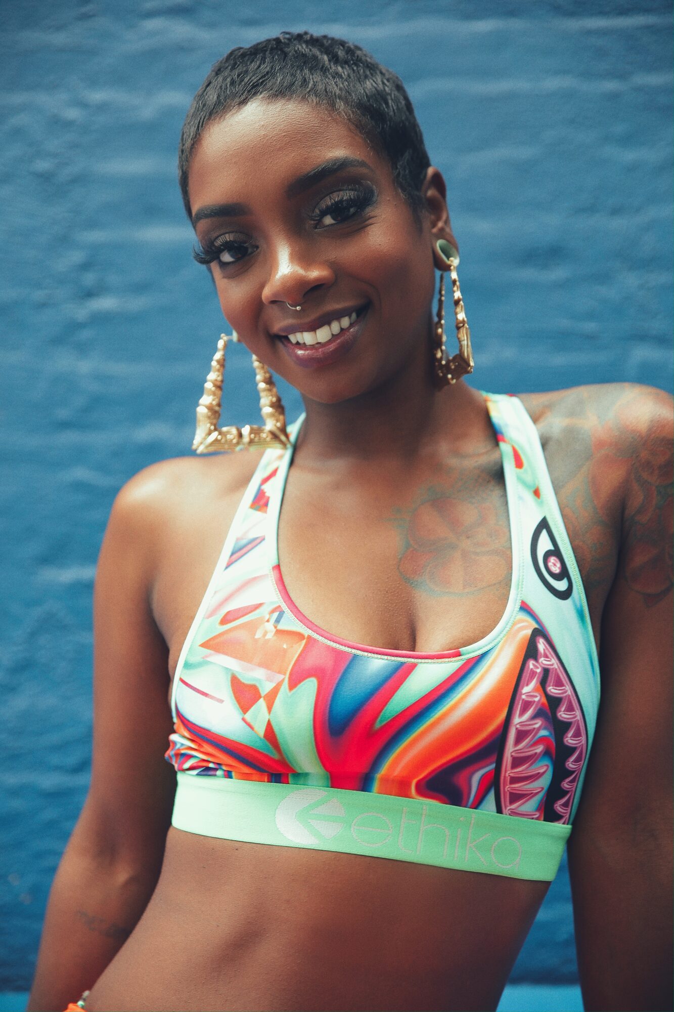 Smiling close-up portrait of a woman with a short pixie cut wearing a colorful abstract-print sports bra against a blue wall.