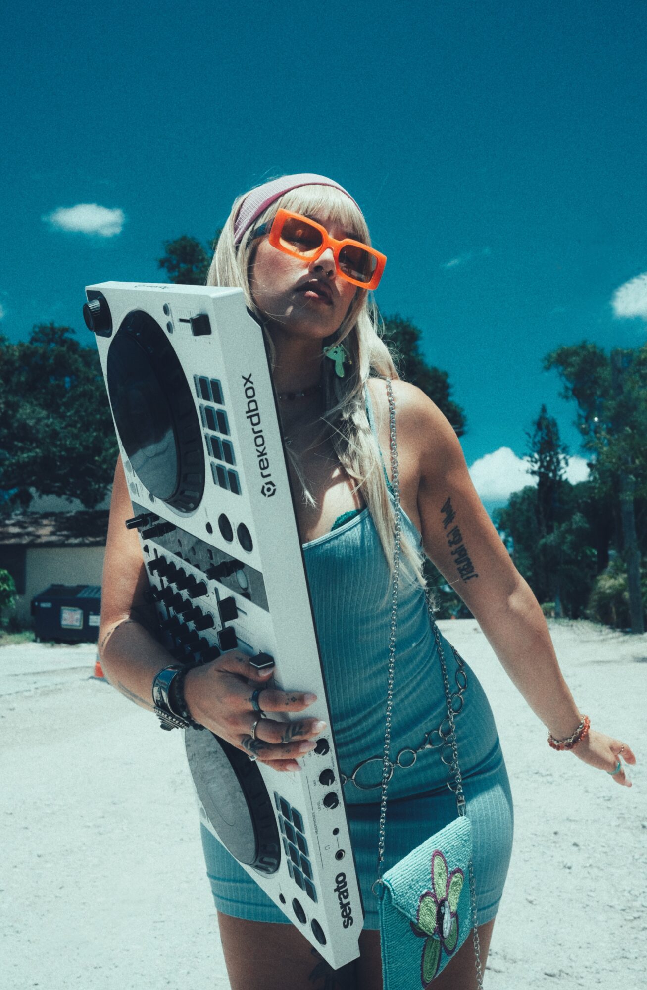 Woman in orange sunglasses and a teal dress holding a DJ turntable controller against a bright blue sky.