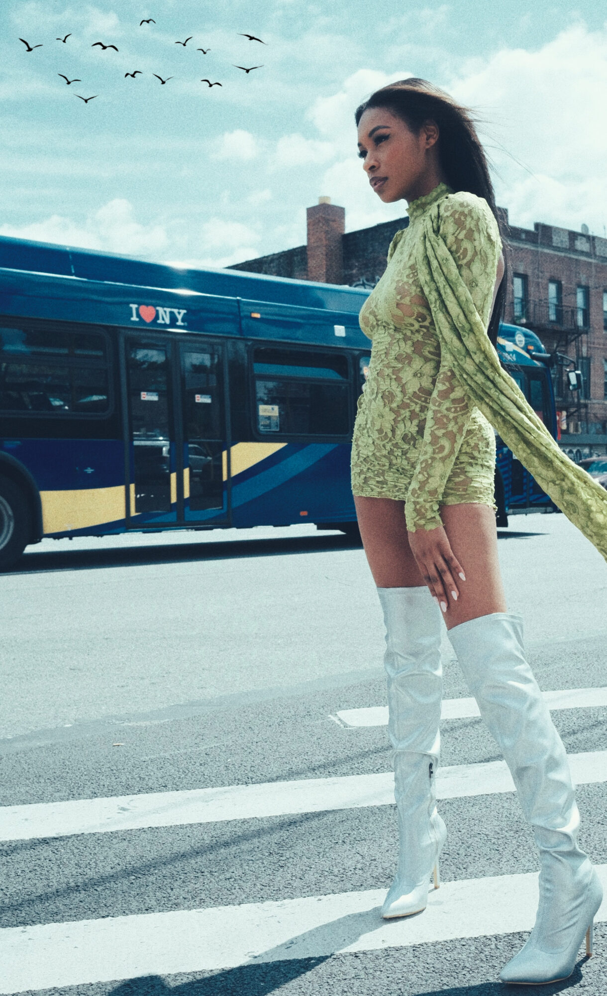 Model in a sheer chartreuse lace dress and white over-the-knee boots crossing a Brooklyn street as an MTA bus passes behind her.