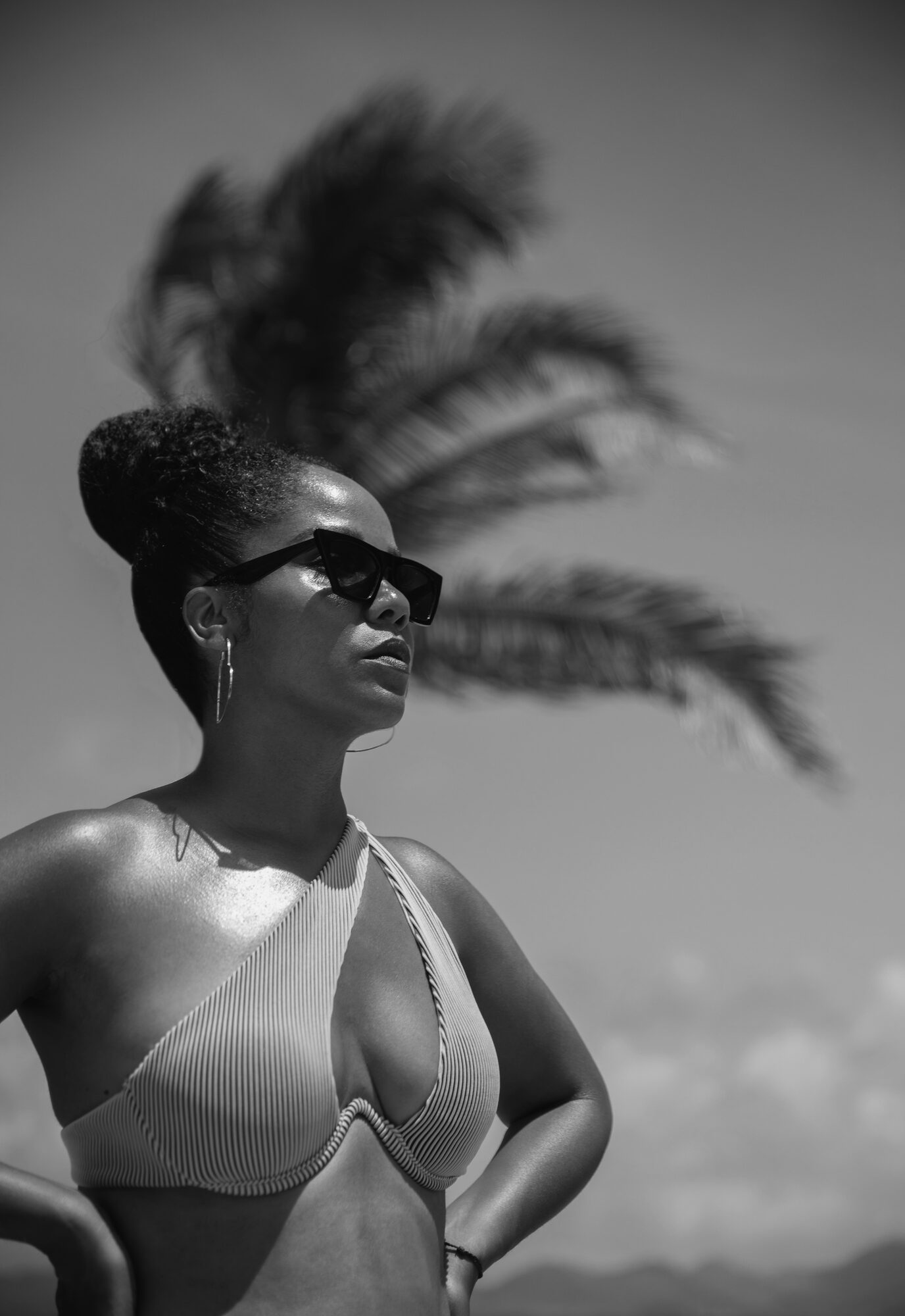 Black and white portrait of a woman in a zebra-print tiered dress leaning on a railing beside a windblown palm tree, ocean and mountains behind her.